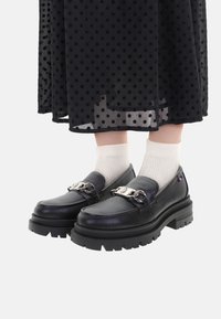 Mocasines de cuero negro con suela gruesa, adornados con una cadena de plata y combinados con calcetas blancas texturizadas. De fondo, se incluye una falda de lunares.