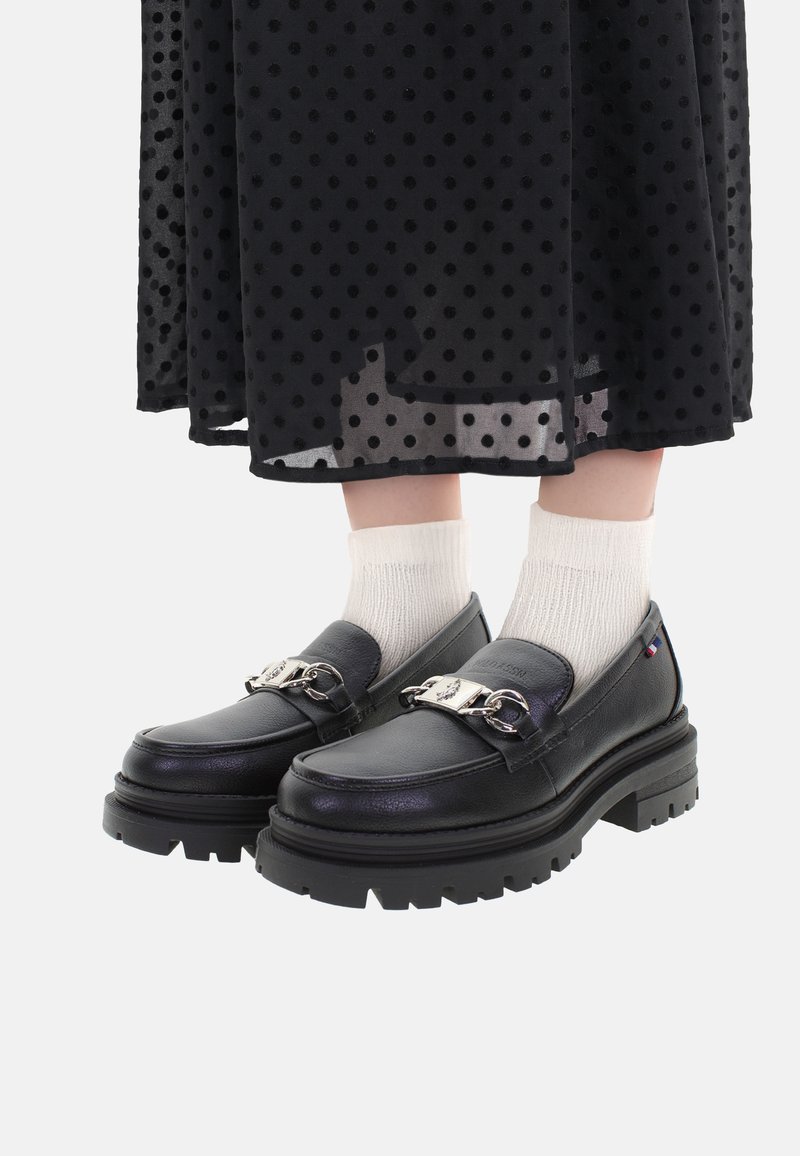 Mocasines de cuero negro con suela gruesa, adornados con una cadena de plata y combinados con calcetas blancas texturizadas. De fondo, se incluye una falda de lunares.