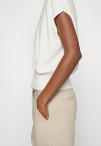 Lehký svetr s krátkým rukávem v barvě slonové kosti, s ribovaným lemem, v kombinaci s béžovými kalhotami na míru, které mají boční kapsu.