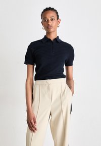 Polo shirt blu navy con maniche corte, motivo testurizzato e bottoni, abbinato a pantaloni beige a vita alta plissettati.