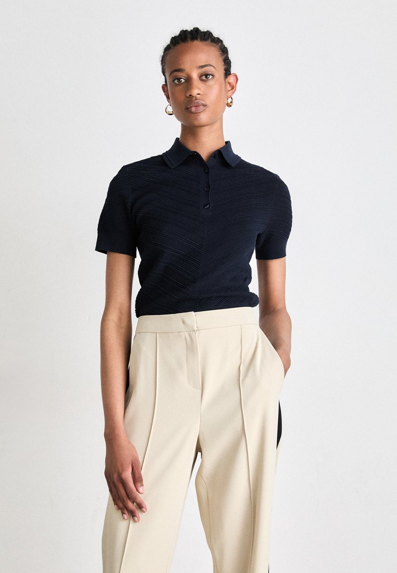 Polo shirt blu navy con maniche corte, motivo testurizzato e bottoni, abbinato a pantaloni beige a vita alta plissettati.