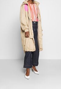 Henkilö, joka on pukeutunut vaaleanbeigeeseen pitkään takkiin, pinkki-oranssiin raidalliseen puseroon, tummiin leveälahkeisiin farkkuihin ja valkoisiin kärkitossuihin, seisoo yksinkertaista taustaa vasten.