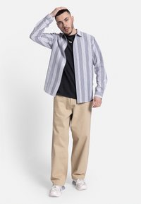 Camisa de botones a rayas en azul y blanco, llevada sobre una camiseta negra, combinada con pantalones de pierna ancha beige y zapatillas de deporte gris claro.
