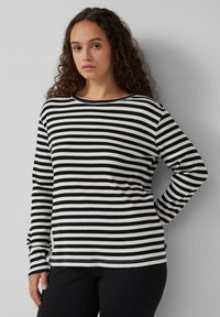 Schwarz-weißer gestreifter Langarmshirt, gerippte Textur, runder Halsausschnitt, lockere Passform, getragen mit dunklen Hosen. Mit einem kleinen Etikett am Saum akzentuiert.