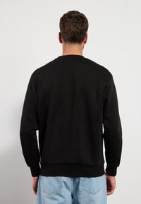 Uomo di spalle con un semplice felpa nera e jeans azzurri contro uno sfondo bianco uniforme.