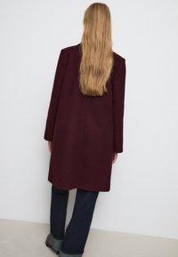Burgunderfarbener Wollmantel mit langen Ärmeln und geradem Schnitt, minimalistischem Nähten und glatter Textur. Kombiniert mit dunklen Jeans.