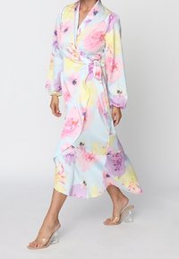 Femme portant une robe portefeuille fleurie pastel à manches longues et des sandales à talons hauts transparentes, marchant devant un fond uni.