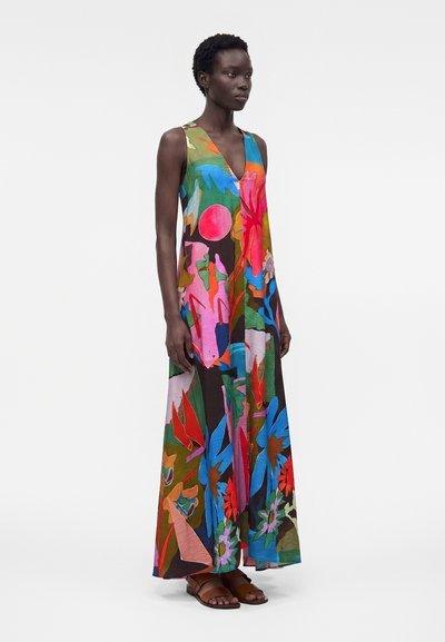 Γυναίκα που στέκεται φορώντας ένα αμάνικο, μακρύ φόρεμα με έντονο αφηρημένο φλοράλ μοτίβο και καφέ φλατ σανδάλια, σε απλό φόντο.