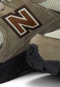 Gros plan sur des baskets en daim beige et mesh, avec un logo "N" marron et blanc cousu, et un détail de semelle en caoutchouc noir texturé.