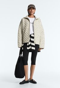 Kobieta w beżowej pikowanej kurtce, białym t-shircie, czarnych legginsach do połowy łydki, czarno-białym paskowanym swetrze związanym w talii, czarnej czapce, niosąca czarną torbę.