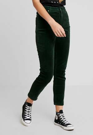 Kvinde iført mørkegrønne bukser i kordfløjl og sorte høje sneakers med hvide snørebånd, stående med det ene ben lidt fremad.