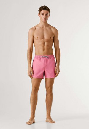 Junger Mann steht barfuß und trägt pinke Badeshorts mit weißem seitlichem Logo vor einfarbigem Hintergrund.