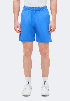 Άνδρας με μπλε αθλητικά σορτς, λευκές κάλτσες και λευκά αθλητικά παπούτσια στέκεται μπροστά σε απλό φόντο.