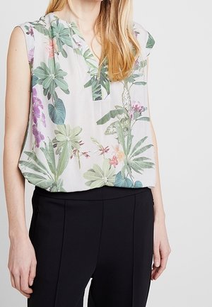 Femme portant un chemisier blanc sans manches avec imprimé floral vert et violet et un pantalon noir sur fond blanc.