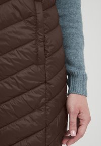 Brun puffervest med chevron-quiltet design, der har en glat tekstur; parret med en lyseblå sweater med ribbet manchetter.