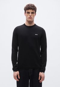 Musta pitkähihainen neulepaita saumattomalla neulekuviolla. Siinä on pyöreä kaula-aukko ja pieni valkoinen "BOSS"-logo edessä.