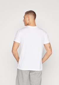 Mand med ryggen vendt, iført en almindelig hvid kortærmet T-shirt og lysegrå bukser, der viser tatoveringer på begge overarme.