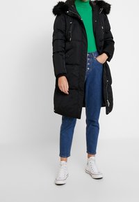 Abrigo negro acolchado con capucha forrada de piel, cierre delantero con cremallera y bolsillos laterales. Combinado con un jersey de cuello alto verde y vaqueros azules de cintura alta.