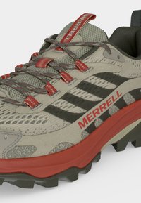 Gros plan d'une chaussure de randonnée Merrell beige et rouge avec une tige en mesh, des accents rouges et une semelle extérieure noire robuste conçue pour une utilisation sur sentiers.