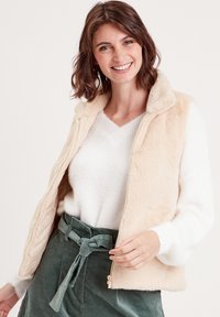 Gilet en fausse fourrure crème avec une fermeture éclair à l'avant, associé à un pull léger et une jupe en velours côtelé verte, avec un détail de taille nouée.