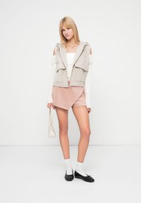 Ανοιχτό γκρι αμάνικο γιλέκο με κουκούλα, λευκό τοπ με ακάλυπτους ώμους, ροζ σορτς από σουέτ με φάσα, μαύρα φλατ παπούτσια και λευκές κάλτσες αστραγάλου. Μικρό τσαντάκι στο χέρι.