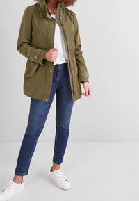Giacca corta verde oliva con cappuccio staccabile, zip frontale, bottoni a pressione, due tasche grandi e vestibilità comoda, indossata sopra una maglietta bianca e jeans blu.