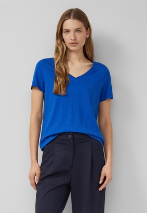 Woman with long blonde hair wearing a bright blue V-neck short sleeve shirt and high-waist dark trousers against a gray background.