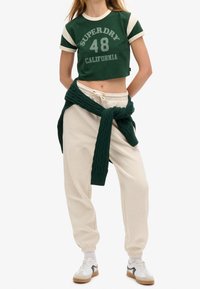 Grøn kortærmet t-shirt med hvide detaljer og trykt "SUPERDRY 48 CALIFORNIA," kombineret med beige sweatpants og en grøn kabelstrikket trøje bundet om taljen.