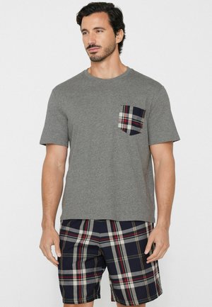 Homme debout portant une chemise grise à manches courtes avec une poche à carreaux et un short assorti à carreaux bleu marine, blanc et rouge sur un fond uni.