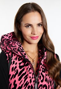 Vest din fâsâlă cu imprimeu de leopard roz, cu glugă, închidere cu fermoar și accente negre. Colier cu pandantiv auriu vizibil, cu o textură netedă a părului.