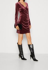 Kort sammetsklänning i vinröd med V-ringning, långa ärmar och draperad design. Kombineras med svarta högskolette stövlar.