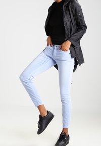 Jeans skinny bleu clair en denim, associés à un haut noir moulant et une veste sombre. Des baskets noires complètent la tenue.