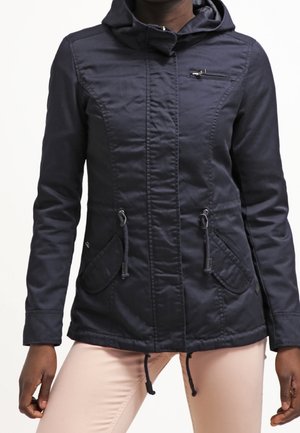 Femme portant une veste à capuche bleu marine foncé avec une fermeture éclair sur le devant et des cordons, associée à un pantalon beige clair sur un fond uni.