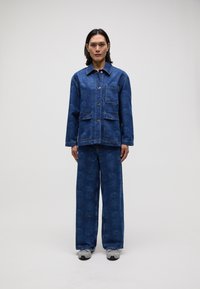 Jeansjakke med et mønster i blått, med frontlommer og knapper. Kombinert med matchende vide bukser og grå sneakers.