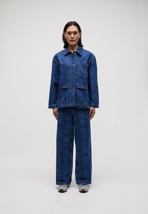 Giacca di jeans con un design a motivi blu, dotata di tasche frontali e bottoni. Abbinata a pantaloni a gamba larga coordinati e sneakers grigie.
