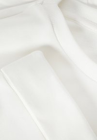 Gros plan sur un tissu en coton blanc montrant une manche pliée et un col côtelé, mettant en valeur la texture et les détails de couture.