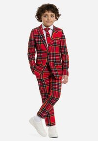 Abito scozzese rosso con accenti neri e gialli, caratterizzato da un blazer con un solo bottone e pantaloni abbinati. Una camicia bianca e una cravatta completano il look.