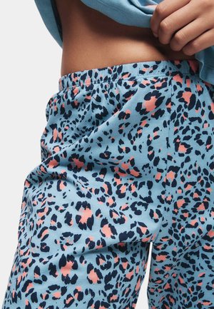 Nahaufnahme einer Person, die die Taille einer blauen Hose mit Leopardenmuster anhebt, wobei der elastische Bund und eine Hand, die ein hellblaues Hemd hält, sichtbar sind.