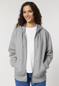 Sivi zip-up dukserica s kapuljačom na žicu, prednjim dzepovima i rebrastim manžetama, nosi se preko bijelog T-shirta i u paru s plavim trapericama.