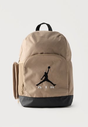 Sac à dos beige et noir avec poche zippée à l'avant présentant un logo silhouette noire de joueur de basket-ball et le texte « AIR » en dessous sur fond uni.