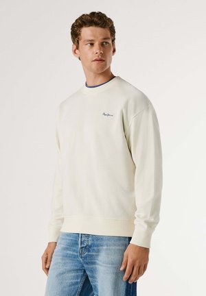 Mand iført hvid langærmet sweatshirt med lille logo på brystet og blå jeans, stående mod en enkel lys baggrund og kiggende mod venstre.