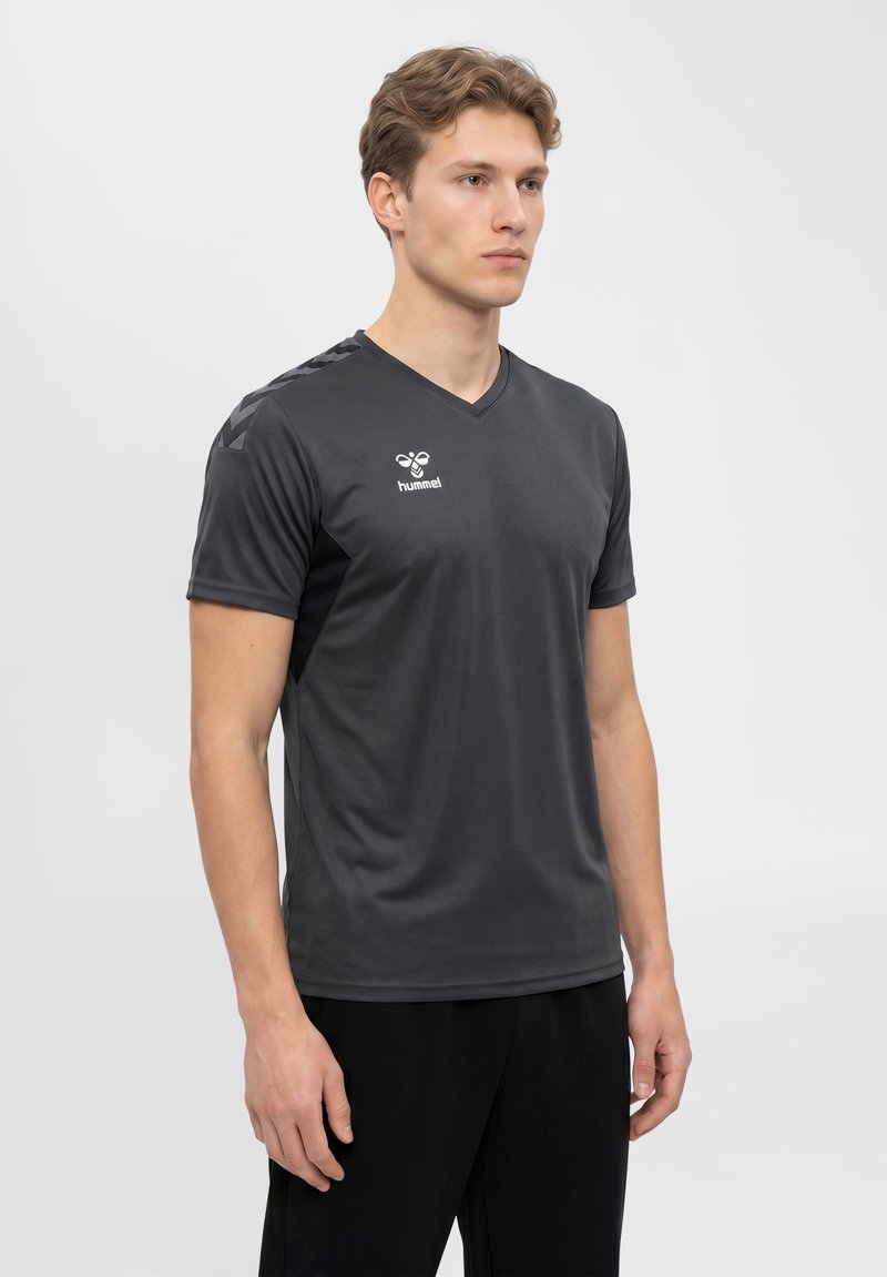 Młody mężczyzna w ciemnoszarej sportowej koszulce z krótkim rękawem z logo hummel na piersi i detalami w kształcie chevron na ramionach.