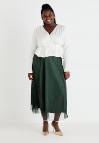 Top peplum bianco a maniche lunghe con scollatura a V, abbinato a una gonna midi plissettata verde fluente e tacchi a punta dorati con dettagli a fibbia.