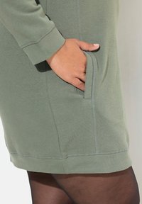El vestido sudadera verde presenta un dobladillo redondeado, puños acanalados, bolsillos laterales y detalles de costura visibles, ofreciendo un aspecto y ajuste relajados.