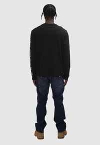 Mann mit geflochtenem Haar, der ein schwarzes Langarmshirt, blaue Jeans und beige Stiefel trägt und mit dem Rücken zu sehen ist vor einfachem Hintergrund.