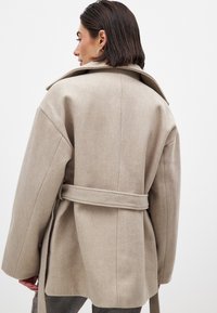 Bež villasegu tõmblukk, millel on lõdvestunud lõige, suur krae, vöökoht ja küljetaskud. Tekstuur tundub pehme ja kerge läikega.