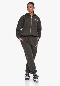 Dunkelbrauner Reißverschluss-Hoodie mit pinkem Logo auf der Brust, kombiniert mit passenden Jogginghosen, beide aus weichem Stoff, getragen mit schwarzen Sneakers.