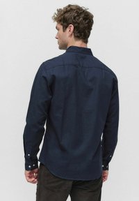 Jonge man met krullend haar, gekleed in een donkerblauwe overhemd met lange mouwen en donkere broek, staande met zijn rug naar de camera tegen een effen achtergrond.