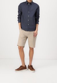 Camicia blu scuro con maniche lunghe, pantaloni corti beige e sneakers marroni. La camicia ha un col formale e bottoni bianchi.