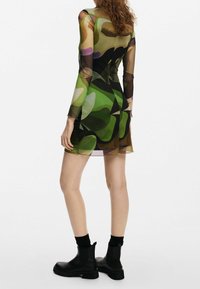 Robe à manches longues avec un motif géométrique transparent multicolore en vert, marron et noir. Portée avec des bottines noires et des chaussettes.